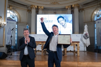 Dos hombres en un escenario. Uno con traje negro y camisa blanca sostiene un diploma enmarcado y levanta el puño. El otro hombre aplaude. Fondo con pantalla y banderas