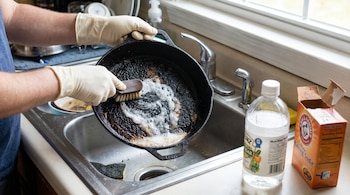 Una persona con guantes lava un sartén quemado en un fregadero, usando un cepillo y productos como bicarbonato de sodio y vinagre.