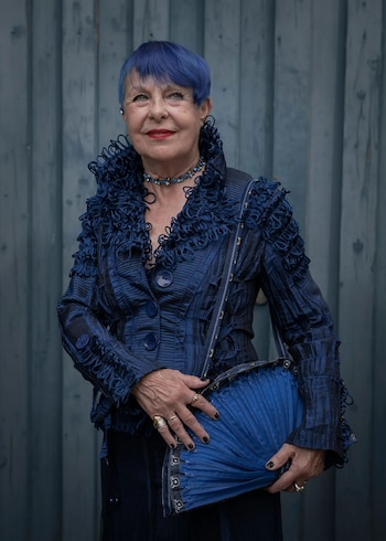 Una mujer de cabello azul corto, chaqueta azul texturizada y labios rojos sonríe frente a una pared azul, sujetando un bolso azul