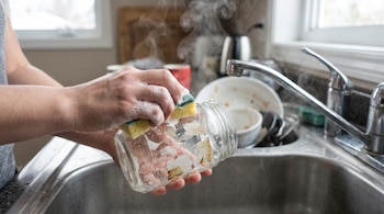 Primer plano de manos lavando un frasco de vidrio con una esponja y espuma en una pileta de cocina. El frasco tiene restos de pegatinas y se ve vapor.