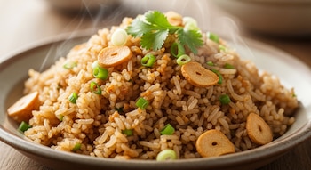 Primer plano de un plato de arroz frito con ajo humeante, decorado con rodajas de ajo dorado, cebolleta verde picada y hojas de cilantro.
