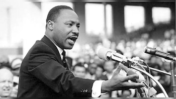 Fotografía en blanco y negro de Martin Luther King Jr. hablando apasionadamente ante un micrófono, con su boca abierta y la mano derecha en el aire