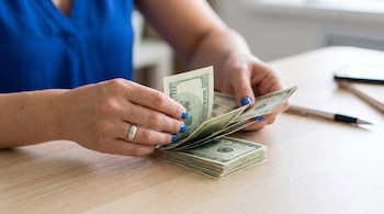 Manos con anillo de plata y uñas azules cuentan varios billetes de cien dólares, que se apilan sobre una mesa de madera clara.