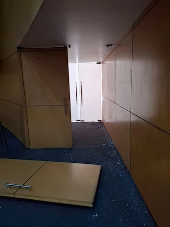Vista interior de un pasillo o aula con paredes de paneles de madera clara y suelo alfombrado azul oscuro con escombros blancos, una puerta abierta y un panel en el suelo