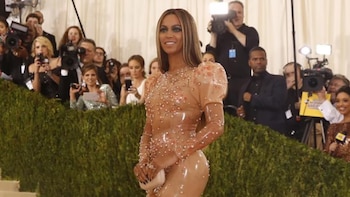 Beyoncé sonríe en una alfombra roja. Viste un vestido color piel brillante con perlas, mangas abullonadas y cola, sobre escaleras y con fotógrafos al fondo
