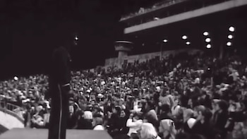 Fotografía en blanco y negro de una persona de espalda sobre un escenario ante una tribuna repleta de espectadores en un estadio