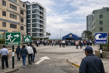 Vista a nivel de calle de personas caminando por una avenida, pasando señales de evacuación, hacia una gran carpa azul. Edificios modernos y antiguos bordean la calle bajo un cielo nublado.