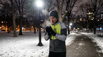 Hombre con gorro, guantes y chaqueta deportiva de invierno en un parque nevado. Vapor sale de su boca bajo una farola encendida.