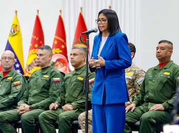 Una mujer con traje azul y lentes habla por micrófono frente a varios hombres en uniforme militar sentados. Hay banderas de Venezuela al fondo