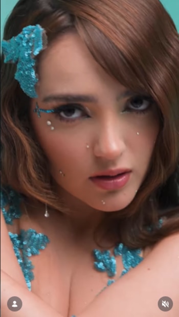 Primer plano de mujer con cabello castaño, maquillaje artístico con delineador azul y gemas blancas, adornos de lentejuelas turquesa en el cabello y cuerpo