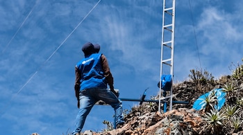 Un hombre de espaldas con chaleco azul de pie sobre rocas junto a una escalera metálica y plantas de montaña bajo el cielo azul.