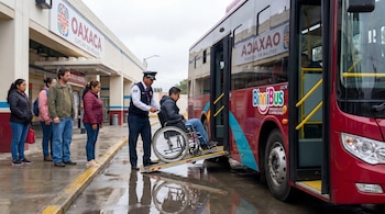 Un conductor de autobús ayuda a un hombre en silla de ruedas a subir a un autobús rojo Binnibus por una rampa en una parada de Oaxaca. Varias personas esperan cerca.