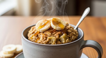 Mug cake de avena y plátano en una taza con una cuchara, decorado con rodajas de plátano, canela y miel, con vapor elevándose.