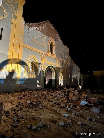 Edificio con fachada gris y amarilla parcialmente colapsada. Escombros de ladrillos y tierra dispersos en el suelo. La toma es nocturna con luces