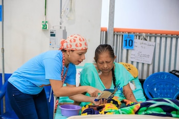 Una profesional de la salud con pañuelo de riñones muestra un folleto a una paciente en cama de hospital. La paciente usa bata verde, con brazo magullado. Equipo médico visible