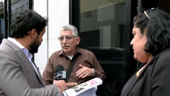 Un reportero sostiene un micrófono frente a un hombre con gafas y una mujer vestida con una chaqueta oscura en la vía pública