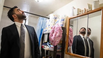 Un hombre barbudo con traje oscuro y corbata mira hacia arriba, con su reflejo en un espejo de tocador; al fondo, artículos de higiene y una manta rosa