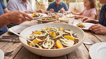 Un tazón grande con ensalada de lentejas, hinojo y gajos de naranja sobre una mesa de madera rodeada por personas comiendo.