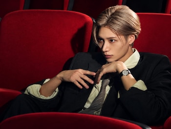 El cantante Taemin sentado en una butaca roja de teatro, viste saco oscuro, camisa clara, corbata y reloj de pulsera