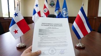 Una mano sostiene un documento oficial frente a pequeñas banderas de Panamá y Costa Rica sobre una mesa de madera.