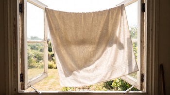 Una toalla blanca grande cuelga de una ventana abierta con el marco de madera descascarado. Al fondo se ve un jardín soleado.