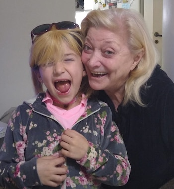 Una mujer rubia y una niña rubia sonríen y posan juntas. La niña guiña un ojo y saca la lengua, llevando gafas de sol en la cabeza y una chaqueta floral