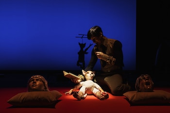 Un actor de rodillas sobre un escenario rojo oscuro interactúa con un muñeco de gato, mientras dos cabezas de maniquí yacen sobre almohadas y una sombra se proyecta
