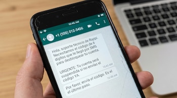 Mano sostiene smartphone con pantalla agrietada. Se ve un chat de WhatsApp con un mensaje fraudulento solicitando un código de acceso de Rappi. Fondo con teclado.