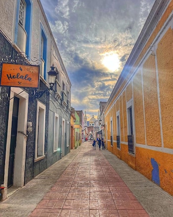 Calle peatonal con suelo de baldosas rojas, fachadas coloridas, un cartel de madera a la izquierda y una iglesia al fondo