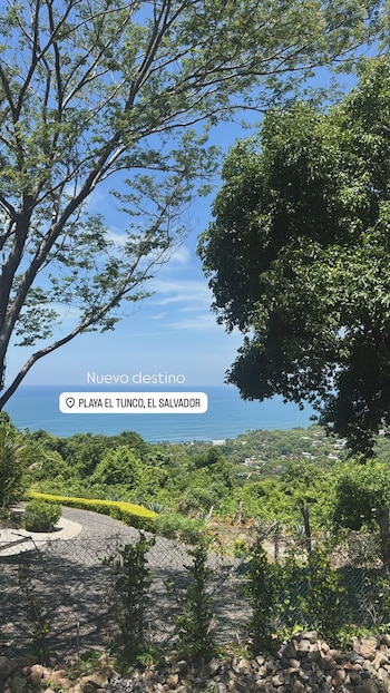 Vista panorámica con árboles, cielo azul, mar, línea costera con edificios, una carretera sinuosa y una valla. Texto: 'Nuevo destino Playa El Tunco, El Salvador'