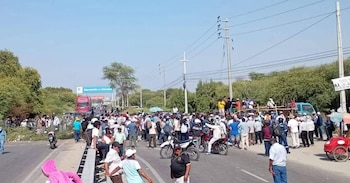 Multitud de personas y varios vehículos, incluyendo un camión rojo y motocicletas, bloquean una carretera rural bajo un cielo despejado. Árboles verdes flanquean la vía