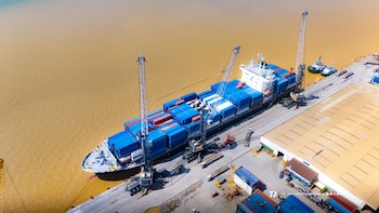 Vista aérea de un buque de carga con contenedores azules y rojos atracado en un muelle gris junto a grúas industriales y agua marrón