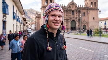 Un hombre joven caucásico sonríe con un chuyo multicolor, gorro andino con orejeras, en una calle adoquinada con personas y una iglesia de fondo.