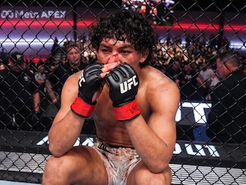 Un peleador con guantes negros de la UFC y manchas de sangre en el rostro permanece sentado detrás de la reja del octágono