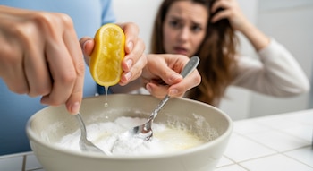Manos exprimen medio limón sobre bicarbonato de sodio en un bol con una cuchara; una mujer se toca el cabello dañado de color castaño, mirando al frente.