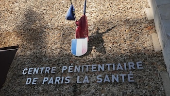 Primer plano de la fachada de piedra de un edificio con la inscripción "CENTRE PÉNITENTIAIRE DE PARIS LA SANTÉ", un escudo francés y una bandera tricolor