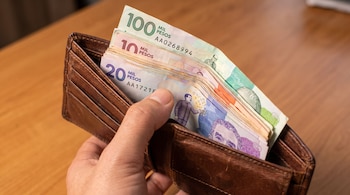 Mano de persona sostiene una cartera de cuero marrón que contiene billetes de pesos colombianos de cien mil, veinte mil y diez mil sobre una mesa de madera clara.