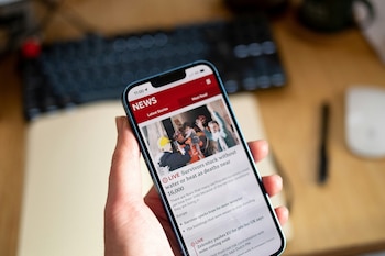 Primer plano de una mano que sostiene un teléfono inteligente con una aplicación de noticias abierta, mostrando titulares e imágenes de rescate. El fondo es un escritorio con un teclado borroso