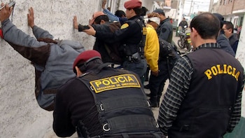 Policías con chalecos y boinas inspeccionan a un grupo de hombres apoyados con las manos sobre una pared blanca