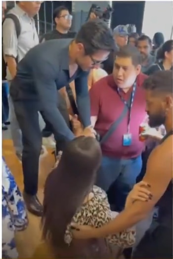 Hombre con camisa oscura y gafas se inclina sobre una persona con cabello castaño largo en una multitud, rodeado de otros asistentes observando la interacción