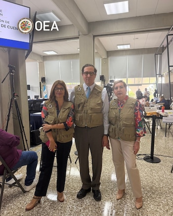Tres observadores con chalecos beige posan de pie en un salón con mesas, computadoras y la bandera de Guatemala al fondo