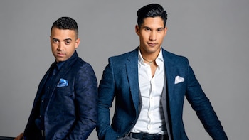 Dos hombres posan frente a un fondo gris; uno con traje estampado azul oscuro, el otro con blazer azul, camisa blanca y collar, ambos mirando al frente