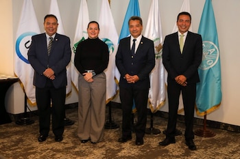 Cuatro personas, tres hombres y una mujer, de pie frente a seis banderas, tres blancas y tres celestes, en un salón interior