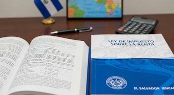 Un manual azul titulado 'LEY DE IMPUESTO SOBRE LA RENTA' y un libro abierto sobre una mesa de madera, con un bolígrafo, calculadora y bandera de El Salvador.