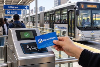 Una mano sostiene una tarjeta azul frente al sensor de un torniquete metálico junto a la vía por donde circula un autobús