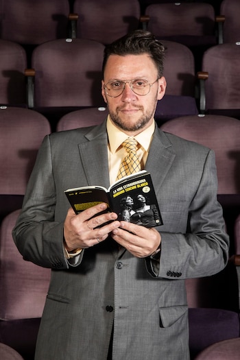 Un hombre con barba y gafas, vestido de traje gris, camisa amarilla y corbata a cuadros, sostiene un libro abierto, mirando a cámara frente a butacas de teatro vacías