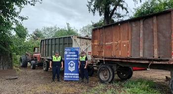 Dos efectivos policiales posan junto a una pancarta azul de la Policía de Tucumán, frente a dos acoplados de gran tamaño y un tractor