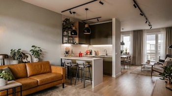 Vista amplia de una sala de estar y cocina de planta abierta con sofá de cuero marrón, barra de cocina con taburetes grises, gabinetes oscuros y piso de madera clara.