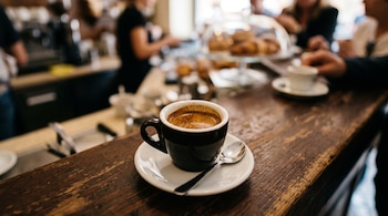 Taza negra con café sobre un plato blanco con una cuchara pequeña en una barra de madera con el fondo desenfocado.