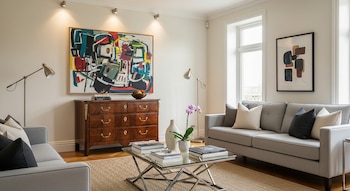 Una sala de estar elegante con dos sofás grises, una cómoda de madera oscura del siglo XVIII, una mesa de centro de cristal y dos obras de arte abstractas en las paredes.